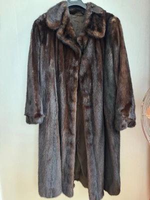 Fantastiskt härlig äkta minkpäls  - Säljer en lyxig lång minkpäls i mörkbrunt med krage, sidofickor och skärp (också i mink). Kappan har en klassisk lätt utställd passform, helfodrad på insidan och osynlig knäppning. Pälsen ger en elegant och tidlös look, samtidigt som den håller dig varm hur kallt det än är.