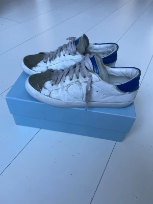 Bud Philippe Model  - Snygga vita Philippe Model sneakers med grå mockadetaljer på tån och blå hälkappa. Klassisk låg modell med vita och grå skosnören. Tillverkade i läder och mocka för en exklusiv känsla. Perfekta för dig som gillar streetstyle och vill ha något unikt. Lägg bud!