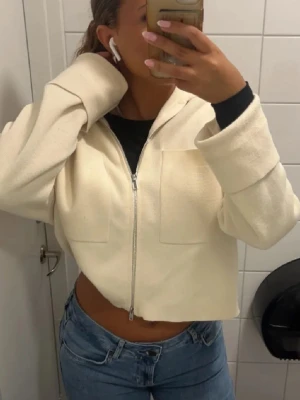 Vit cropped hoodie med dragkedja - Säljer en vit cropped hoodie med dragkedja framtill och två stora bröstfickor. Hoodien har en avslappnad passform och är tillverkad i mjukt material. Perfekt att matcha med jeans för en chill look. Kan användas som vårjacka😊