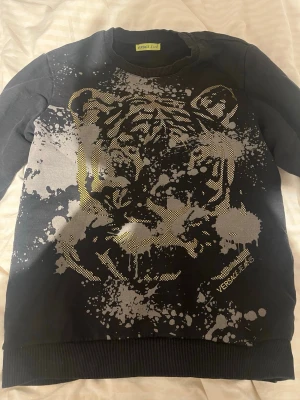  Versace Jeans tröja - Cool svart sweatshirt från Versace Jeans med stort tigertryck i silver och gult framtill. Tröjan har rund halsringning och ribbade muddar. Materialet ser ut att vara bomullsmix. Perfekt för dig som gillar statement-plagg och streetwear.
