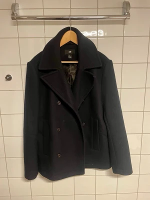 Marinblå skepparkavaj från H&M - Stilren marinblå skepparkavaj från H&M med dubbelknäppning och bred krage. Klassisk modell med två sidofickor och slät baksida. Tillverkad i ett tjockare tyg som ger en snygg siluett och passar perfekt till höst och vinter.