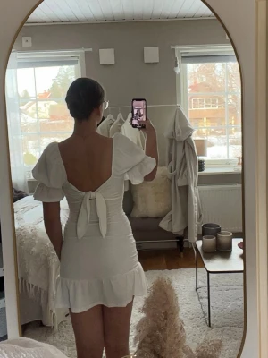 Vit kort klänning med volang och knyt - Supersöt vit kort klänning med puffärmar. Den har en knytning som är till för att ha framåt, men är även supersnygg att ha bak- och- fram. Klänningen har volangkant nertill och är figurnära i modellen, samtidigt som den är väldigt stretchig och bekväm. Passar både XS, S och säkert M. Det går att anpassa klänningens längd som ni ser på bild 1 och 2, samma gäller puffärmen. Perfekt till studenten eller sommaren! Använd en gång på skolavslutning. Säljer eftersom den inte kommer till användning🌸