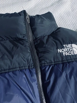 Blå dunjacka från The North Face - Snygg blå dunjacka från The North Face med svart axelparti, hög krage och dragkedja framtill. Jackan har två framfickor med dragkedja och broderad logga på bröstet. Perfekt för kalla vinterdagar och riktigt trendig stil.