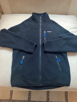 Mörkblå teddyjacka från Patagonia - Mysig mörkblå teddyjacka från Patagonia med hel dragkedja framtill och tre fickor med blå detaljer på dragkedjorna. Jackan har hög krage och är tillverkad i fluffigt teddy-material som håller dig varm under kyliga dagar.