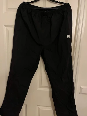 Svarta täckbyxor Helly Hansen - Säljer ett par svarta regnbyxor från Helly Hansen med elastisk midja och meshfoder. Perfekta för kalla dagar och outdoor-äventyr. Byxorna har en enkel och stilren design med HH-logga på låret.