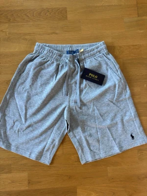 Grå shorts från Polo Ralph Lauren - Snygga grå shorts från Polo Ralph Lauren med klassisk logga broderad i svart på benet. Shortsen har dragsko i midjan och är tillverkade i mjuk bomull, perfekta för en avslappnad stil. Två sidofickor och en bekväm passform.