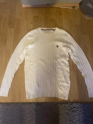 Vit kabelstickad US polo tröja - Kabelstickad vit tröja med rund halsringning och lång ärm. Tröjan har ett diskret broderat emblem på bröstet och ribbade muddar vid ärmslut och nederkant. Perfekt för lager på lager och enkel att matcha med olika outfits.