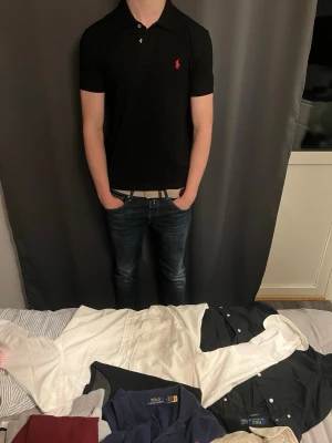Svart pikétröja från Polo Ralph Lauren - Klassisk svart pikétröja från Polo Ralph Lauren med röd broderad logga på bröstet. Tröjan har korta ärmar, krage och knappar framtill. Perfekt till jeans för en clean och stilren look.