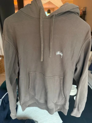 Brun hoodie från Stüssy med logga - Säljer en brun hoodie från Stüssy med vit broderad logga på bröstet. Hoodien har huva med dragsko och en stor magficka. Tillverkad i mjukt bomullsmaterial och har en avslappnad passform som är perfekt för en chill stil.