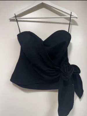 Svart bandeu topp med rosett - Trendig svart bandeu topp stor rosettdetalj i sidan. Toppen har en snygg dragkedja i ryggen och en croppad passform som ger en modern look. Perfekt för dig som vill sticka ut med en stilren och cool topp. Storlek M men passar S