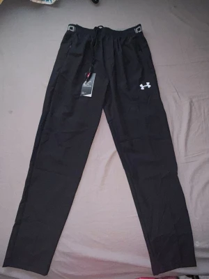 Svarta träningsbyxor från Under Armour - Säljer ett par svarta träningsbyxor från Under Armour med dragsko i midjan och logga på vänster ben. Byxorna har elastisk midja med textdetalj och är tillverkade i ett lätt och funktionellt material som passar perfekt till träning.