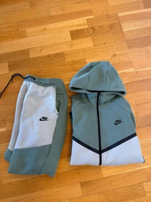 Nike tech fleece tracksuit (tröja+byxor) - Nike tech fleeve set i mjuk bomullsblandning. Setet består av en hoodie med dragkedja och huva samt matchande byxor. Färgen är en mix av ljusgrönt och vitt med svarta detaljer och Nike-logga på både tröja och byxor. Perfekt för chill dagar hemma.