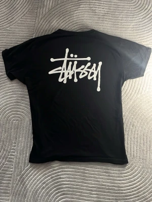 Stussy Tröja - Tja! Säljer en svart basic Stüssy t-shirt med tryck på frambröstet och på ryggen. Tröjan har inga defekter och är i väldigt fint skick! Passformen är najs och den har skönt material. Perfekt till sommarväder och funkar lika bra att ha under någon annan tröja. Hör av er om frågor finns, tack i förväg!