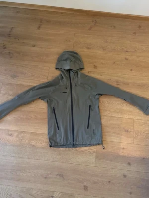 Grå Gore-Tex Jacka från Mammut - Snygg olivgrön regnjacka från Mammut med huva och hel dragkedja framtill. Två sidofickor med dragkedja och justerbar nederkant. Tillverkad i lätt och vattenavvisande material, perfekt för blöta dagar.