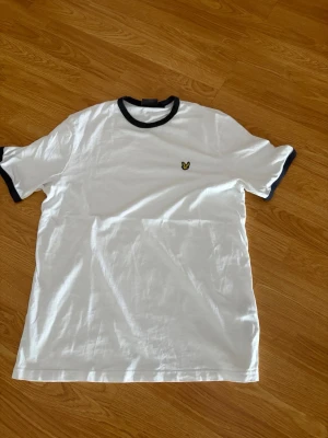Vit t-shirt från Lyle & Scott - Vit t-shirt från Lyle & Scott med svarta detaljer vid krage och ärmslut. Klassisk passform och liten gul logga på bröstet. Perfekt för en clean och stilren look.