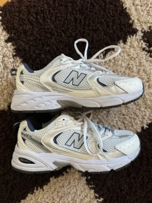 New Balance 530 vita sneakers - Snygga New Balance 530 sneakers i vitt med silver- och marinblå detaljer. Ovandelen är i mesh och syntetmaterial för en sportig look. Klassisk N-logga på sidan och ABZORB-dämpning i sulan för extra komfort. Perfekta för en trendig och avslappnad stil.