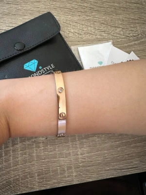 Rosefärgad armband med stenar från Diamond style - Snyggt armband i rosefärg från Diamond Style London. Armbandet är stelt och dekorerat med små gnistrande stenar och skruvliknande detaljer runt om. Perfekt accessoar för att lyfta din outfit. Helt oanvänd och armbandet är 16,5cm i omkrets! OBS: FRAKTAR INOM 24H!🩷 