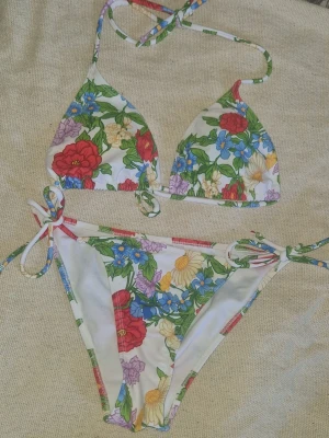 Bikini med knyt - Säljer en färgglad trekantsbikini med blommigt mönster. Både överdel och underdel har knytband i sidorna och nacken för justerbar passform. Perfekt för dagar på stranden eller poolen. Aldrig använt då jag istället använder mina andra, tyvärr har hygienskyddet på nedre delen lossnat. 