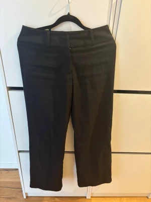 Svarta raka kostymbyxor - Snygga svarta kostymbyxor med rak passform och klassiska pressveck. Byxorna har hällor för skärp och stängs med knapp och dragkedja. Perfekta för en stilren och clean look. Materialet ser ut att vara en blandning av polyester och viskos.