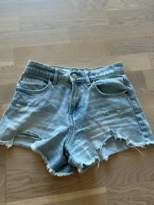 Ljusblåa jeansshorts med fransig kant - Säljer ett par ljusblåa jeansshorts med fransig, rå kant och slitna detaljer. Shortsen har fem fickor, bälteshällor och knappgylf. Perfekta för varma dagar och ger en avslappnad vibe.