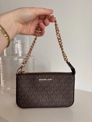 Brun  Michael Kors - Snygg brun väska från Michael Kors med guldfärgad kedjerem och dragkedja. Väskan har ett diskret MK-mönster över hela ytan och loggan i guld framtill. Tillverkad i tåligt konstläder och perfekt storlek för det viktigaste. Nypris 1600