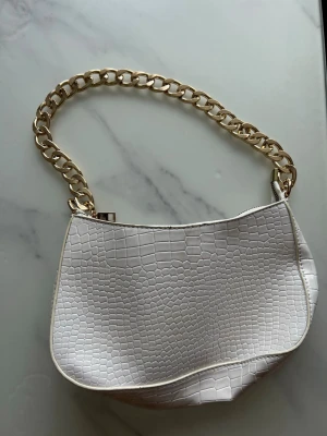 Vit axelväska med kedja - Snygg vit handväska med croco-mönstrad yta och en chunky guldfärgad kedja som axelrem. Väskan har en modern, böljande form och stängs med dragkedja. Perfekt för att lyfta vilken outfit som helst.