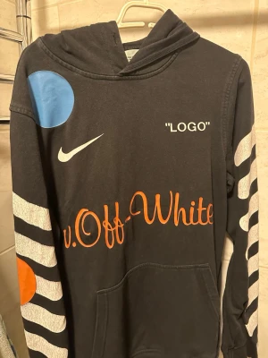 Svart off white hoodie  - Cool svart hoodie med huva, stora vita och orange tryck på både fram- och baksida samt armar. Har texten 'LOGO', Nike-logga och 'Off-White' i orange. Färgdetaljer i blått och orange på axel och ärm. Känguruficka framtill och randiga vita detaljer på ärmarna.