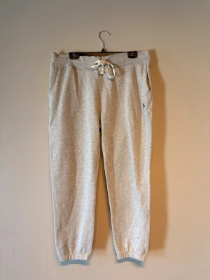 Grå mjukisbyxor från Polo Ralph Lauren - Snygga grå mjukisbyxor från Polo Ralph Lauren med dragsko i midjan och resår vid bensluten. Byxorna har sidofickor och den klassiska broderade loggan i mörkblått på ena benet. Perfekta för en chill och avslappnad stil.