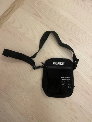Svart axelväska från Hoodrich - Snygg svart axelväska från Hoodrich med justerbar rem och flera fack. Väskan har vit logga och text framtill samt dragkedja. Perfekt för att bära det viktigaste och ger en streetwear vibe till din outfit.