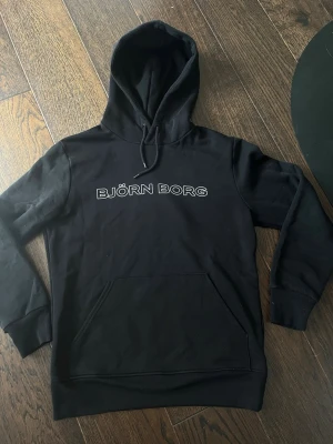 Björn Borg hoodie  - Säljer en svart hoodie från Björn Borg med stor logga i vitt på bröstet. Hoodien har huva med dragsko och en stor magficka framtill. Perfekt för en chill och sportig look. Tillverkad i mjukt material och har ribbade muddar vid ärmar och nederkant.