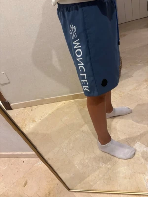 Blå shorts med tryck från Moncler - Säljer ett par blå shorts från Moncler med vitt tryck längs sidan och svart Moncler-logga. Shortsen har snörning i midjan och är tillverkade i ett lätt syntetmaterial, perfekta för sommaren.