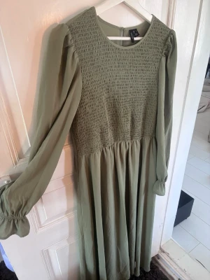 Grön långärmad klänning från H&M - Snygg grön klänning med lång ärm och smockad överdel. Klänningen har rund halsringning och ett mjukt, flytande tyg som ger en avslappnad men ändå chic look. Perfekt för dig som gillar stilrena detaljer och en bekväm passform.