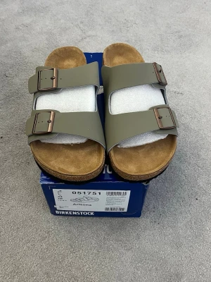 Gröna Birkenstock Arizona sandaler - Snygga Birkenstock Arizona sandaler i olivgrönt med två breda remmar och justerbara metallspännen. Fotbädden är i mjukt mocka och sulan är i kork för extra komfort. Perfekta för en avslappnad och trendig stil.