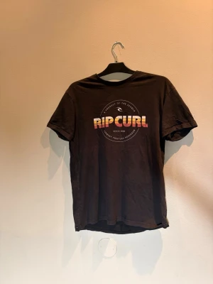 Svart Rip Curl t-shirt med tryck - Snygg svart t-shirt från Rip Curl med färgglatt tryck framtill i gult, orange och rött. Klassisk rund hals och korta ärmar. Perfekt för dig som gillar surfvibbar och streetstyle.