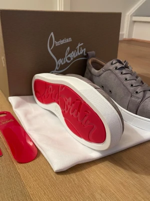 Gråa sneakers från Christian Louboutin - Stilrena gråa sneakers i mocka från Christian Louboutin med klassisk röd sula och vita kanter. Skorna har rund tå, gråa skosnören och diskreta sömdetaljer. Perfekta för dig som vill ha en lyxig och trendig look med ikonisk design.