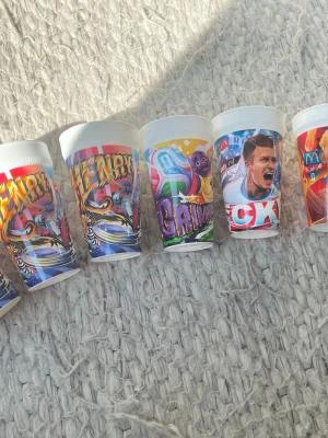 7 st Samlings muggar - Säljer ett gäng plastmuggar med olika färgglada motiv och tryck, bland annat sport och tecknade figurer. Muggarna är vita i grunden och har tryck i rött, blått, gult, lila och grönt. Perfekta till samling eller temafest. Eftertraktade muggar