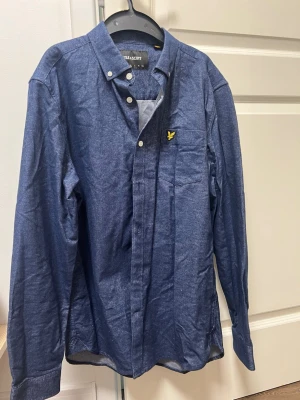 Blå skjorta från Lyle & Scott - Snygg blå skjorta från Lyle & Scott med klassisk krage och knappstängning. Skjortan har lång ärm och en liten gul logga på bröstfickan. Perfekt för en stilren look och passar till många tillfällen.