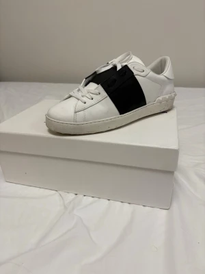 Valentino sneakers - Snygga vita sneakers från Valentino med bred svart rem över vristen och klassisk snörning. Skorna har en stilren och modern design med låg profil och är tillverkade i slätt läder. Perfekta för dig som gillar minimalistisk stil och vill ha något som sticker ut.