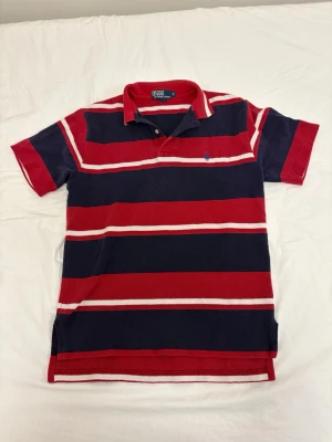 Vintage Ralph Lauren piké - Klassisk pikétröja från Polo Ralph Lauren i rött, marinblått och vitt. Tröjan har breda ränder, krage och knappar vid halsen samt den ikoniska broderade loggan på bröstet. Tillverkad i bomull och perfekt för en avslappnad men stilsäker look.