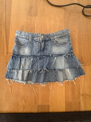 Blå jeanskjol med volanger - Kort jeanskjol i blå denim med två lager volanger och fransiga kanter. Kjolen har klassiska fickor fram  samt hällor för bälte.