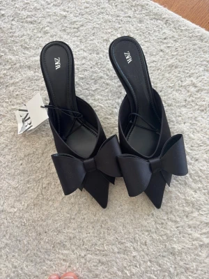 Svarta pumps med rosett från Zara - Stilrena svarta pumps från Zara med spetsig tå och stor dekorativ rosett framtill. Skorna har öppen häl och låg klack, perfekta för att lyfta din outfit med en trendig touch. Materialet är slätt och ger en elegant look.