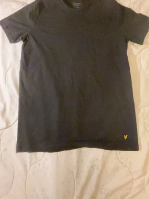 Svart Lyle&Scott-shirt med gul broderad logga - En stilren Lyle&Scott svart t-shirt i bomull med en gul broderad logga på nedre delen av framsidan. Klassisk passform och rund halsringning, perfekt till jeans eller shorts. Enkel men snygg design som funkar till det mesta.