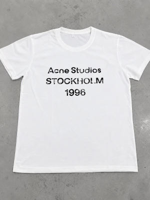 Vit Acne Studios t-shirt - Säljer en vit t-shirt från Acne Studios med svart text 'STOCKHOLM 1996' på bröstet. Klassisk passform och rund halsringning. Perfekt basic-plagg med stilren design och mjukt bomullsmaterial.