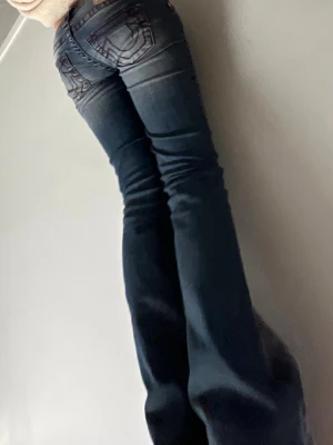 Mörkblå bootcut jeans från true religion - Säljer ett par mörkblå bootcut jeans med snygga broderade detaljer på bakfickorna i vin röd. Jeansen har låg midja. Ber om ursäkt för den dåliga bilden men det va den jag hade så man ser inte jätte bra
