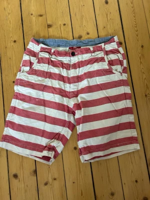 Randiga shorts från Peanloco - Säljer ett par snygga shorts från Peanloco i rött och vitt med breda horisontella ränder. Klassisk passform med fickor fram och bak, knapp och dragkedja. Perfekta för sommaren och tillverkade i mjuk bomull.