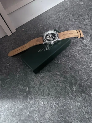 Silverfärgad klocka med läderband - Stilren klocka med silverfärgat boett och svart urtavla med flera små ur. Armbandet är i ljust brunt läder med texten 'poedagar' ingraverad. Kommer med originalförpackning och verktyg för bandbyte.