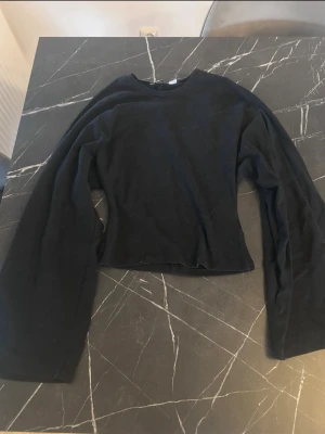 Svart långärmad tröja med vida ärmar - En svart långärmad tröja med rund hals och vida ärmar. Tröjan har en enkel, minimalistisk design och är något croppad i modellen. Perfekt för dig som gillar en avslappnad och stilren look.