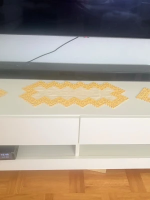 Hardanger duk Gul spetsduk med blommönster - Säljer en rektangulär duk i vitt tyg med gul spetskant och blommönster. Duken har ett sicksackmönster längs kanten och mäter cirka 44 cm på längden. Perfekt som dekoration på bord eller tv-bänk.