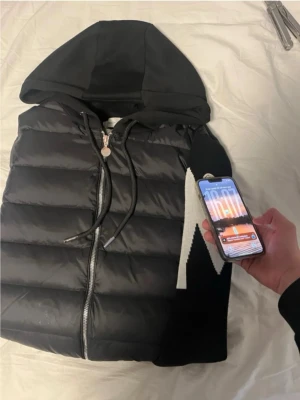Moncler Cardigan  - Snygg svart dunväst med huva och långa stickade ärmar. Västen har dragkedja framtill, dragsko i huvan och ribbade muddar. Moncler-logga på ena ärmen och quiltad design på framsidan ger en sportig och trendig look. Pris går att diskutera vid snabbaffär och NFC-tag funkar.