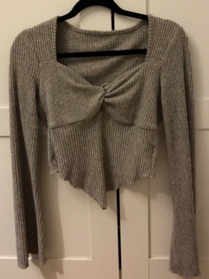 Grå ribbad topp med twist framtill - Trendig grå ribbad topp med lång ärm och twistad detalj framtill. Toppen har en asymmetrisk nederkant och fyrkantig ringning som ger en unik look. Perfekt för dig som vill ha något extra till din outfit.